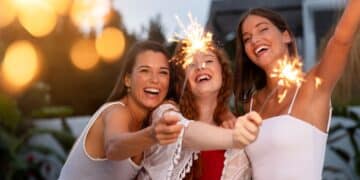 Três amigas sorridentes celebrando ao ar livre durante o entardecer, segurando velas faíscas (sparklers) acesas que criam pontos de luz brilhantes.