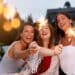 Três amigas sorridentes celebrando ao ar livre durante o entardecer, segurando velas faíscas (sparklers) acesas que criam pontos de luz brilhantes.