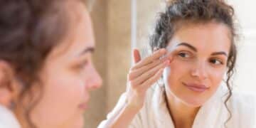 Mulher jovem com pele radiante apontando para a área dos olhos, demonstrando firmeza e hidratação na pálpebra inferior.