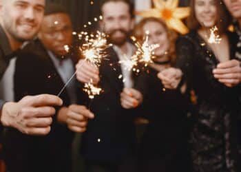 Grupo de amigos multiculturais vestidos com roupas de festa escuras, sorrindo e segurando velas faíscas acesas (sparklers) em um ambiente festivo com luzes douradas ao fundo.