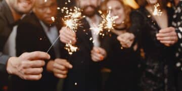 Grupo de amigos multiculturais vestidos com roupas de festa escuras, sorrindo e segurando velas faíscas acesas (sparklers) em um ambiente festivo com luzes douradas ao fundo.