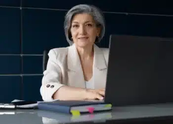 Idosa sorrindo em ambiente de escritório com computador, em referência à isenção de taxas de conselhos profissionais para idosos com mais de 20 anos de experiência.