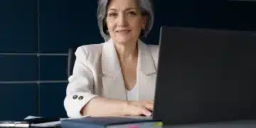 Idosa sorrindo em ambiente de escritório com computador, em referência à isenção de taxas de conselhos profissionais para idosos com mais de 20 anos de experiência.