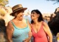 Duas mulheres idosas sorrindo e conversando na praia; uma usa maiô listrado e chapéu de palha, e a outra usa maiô coral, exemplificando moda praia confortável.