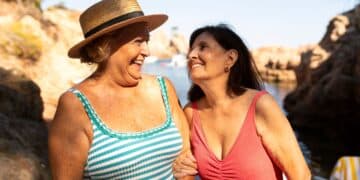 Duas mulheres idosas sorrindo e conversando na praia; uma usa maiô listrado e chapéu de palha, e a outra usa maiô coral, exemplificando moda praia confortável.