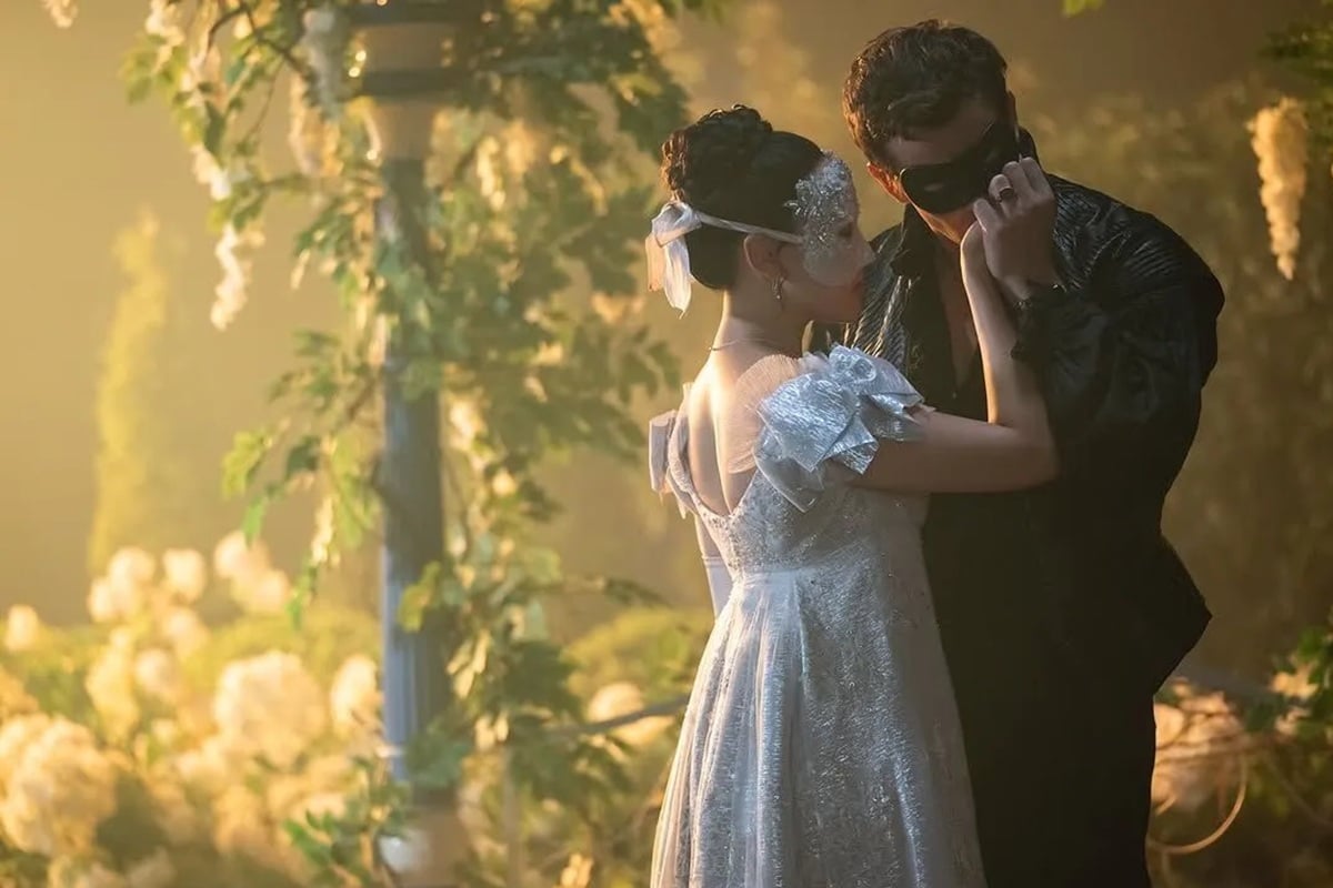 Casal elegante usando máscaras, vestido branco e traje preto, em dança íntima com iluminação suave e flores ao redor