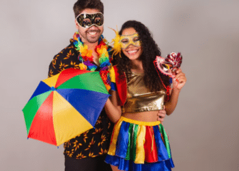 Casal jovem sorridente vestindo fantasias coloridas de Carnaval com máscaras douradas e pretas, segurando uma sombrinha de frevo e colar de flores.