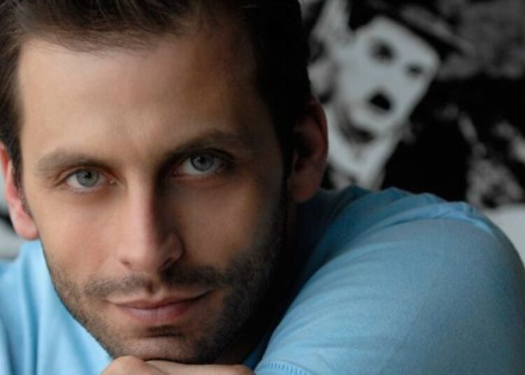 Close do rosto do ator Henri Castelli, com olhos azuis em destaque, barba rala e expressão serena. Ele está apoiando o queixo sobre a mão, vestindo uma camiseta azul clara.