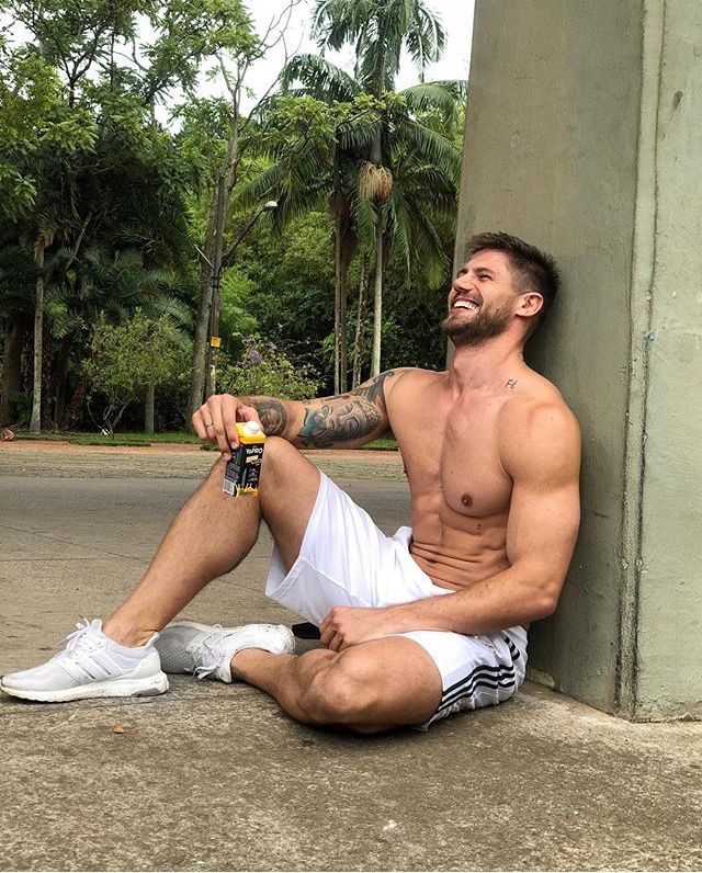 Fotografia de Jonas Sulzbach, modelo e influenciador gaúcho, sentado no chão encostado em uma pilastra, sorrindo e segurando uma bebida esportiva em um parque.
