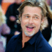 Close do ator Brad Pitt sorrindo e olhando para o lado, vestindo uma camisa azul escura, em um ambiente externo com pessoas ao fundo.