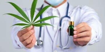 Médico com jaleco branco, estetoscópio e máscara cirúrgica azul, estendendo as mãos para a câmera. Na mão esquerda, ele segura uma folha verde de cannabis, e na mão direita, um pequeno frasco conta-gotas verde-escuro com tampa dourada, simbolizando o óleo medicinal.