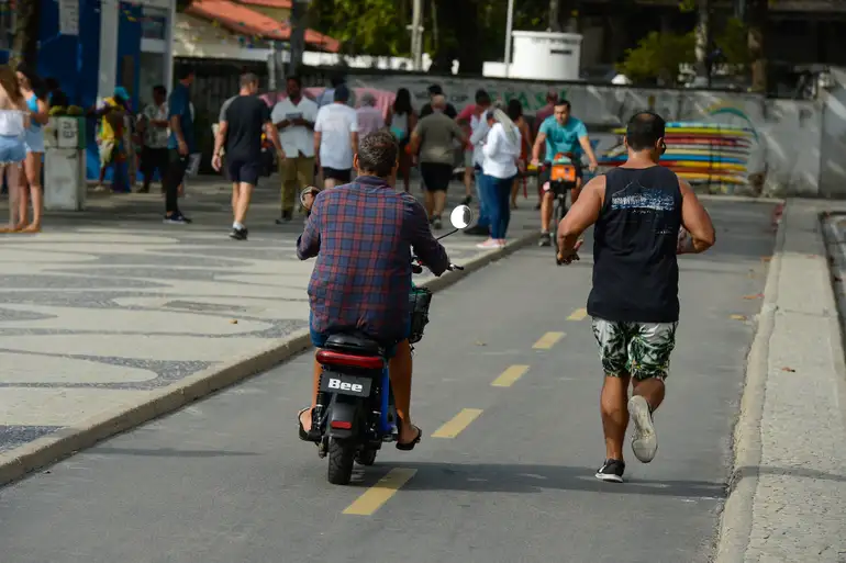 Ciclomotores e as novas regras de trânsito Pessoa em ciclomotor no meio de ciclistas e pedestres, destacando a necessidade de registro, placa e CNH, conforme as novas regras.