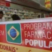 Cartaz do Programa Farmácia Popular em farmácia com prateleiras e atendentes ao fundo
