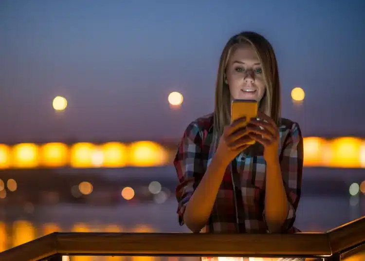 Mulher sorrindo e usando celular à noite, com luzes suaves ao fundo, ideal para compartilhar mensagens inspiradoras.