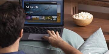 Homem sentado no sofá mexendo no notebook assistindo documentário Shark Friends com tigela de pipoca ao lado