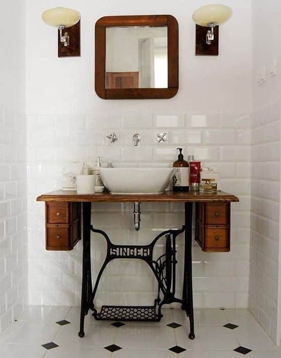 Base de ferro de uma máquina de costura Singer antiga adaptada como bancada de lavabo, com tampo de madeira rústica, cuba branca e espelho sobre parede de azulejos brancos.