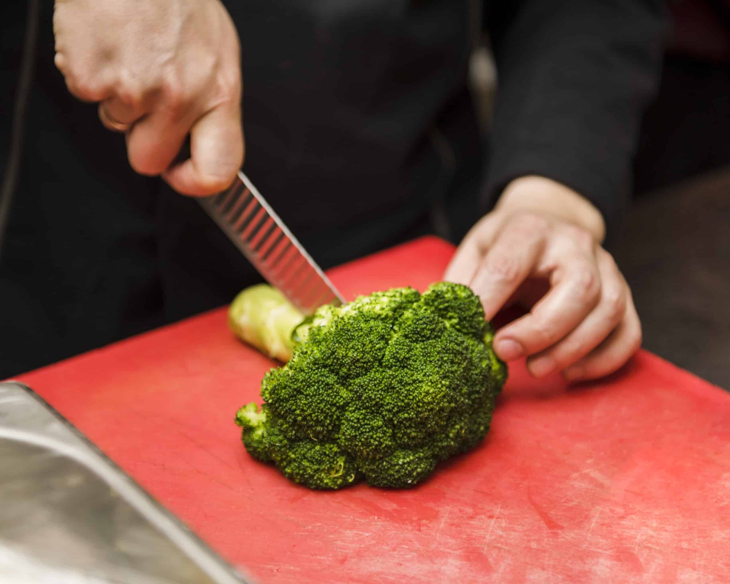 Close-up das mãos de uma pessoa utilizando uma faca para cortar floretes de brócolis fresco sobre uma tábua de corte vermelha em uma cozinha.