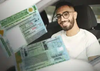 Homem com barba e óculos sorrindo sentado no carro segurando carteira de motorista brasileira