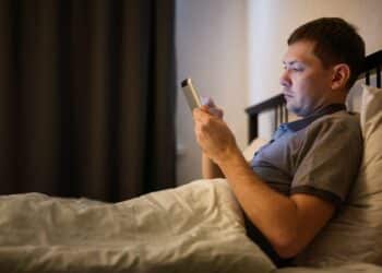 Homem deitado na cama à noite, em um quarto escuro, segurando um tablet iluminando seu rosto. Ele demonstra expressão séria enquanto usa o dispositivo antes de dormir.