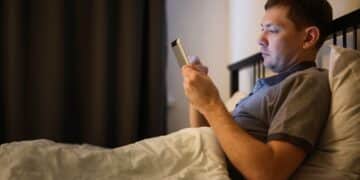 Homem deitado na cama à noite, em um quarto escuro, segurando um tablet iluminando seu rosto. Ele demonstra expressão séria enquanto usa o dispositivo antes de dormir.