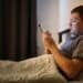 Homem deitado na cama à noite, em um quarto escuro, segurando um tablet iluminando seu rosto. Ele demonstra expressão séria enquanto usa o dispositivo antes de dormir.