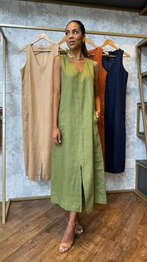 Mulher sorridente com pele morena e cabelos presos, posando em uma loja de roupas, vestindo um vestido midi verde-oliva sem mangas com fenda frontal e sandálias de tira bege, enquanto outros vestidos de linho em tons terrosos estão pendurados ao fundo.