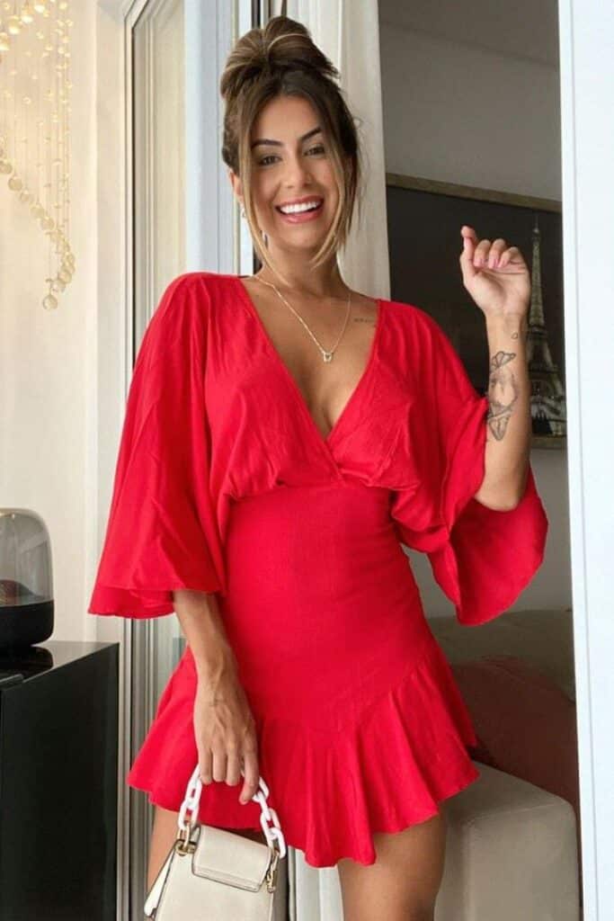 Mulher sorridente com cabelos presos em coque e maquiagem destacada, posando em um ambiente interno, vestindo um vestido vermelho curto com mangas amplas, decote em V e saia com babados, segurando uma pequena bolsa branca.