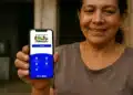 Mulher sorridente mostrando smartphone com aplicativo Bolsa Família em frente à casa