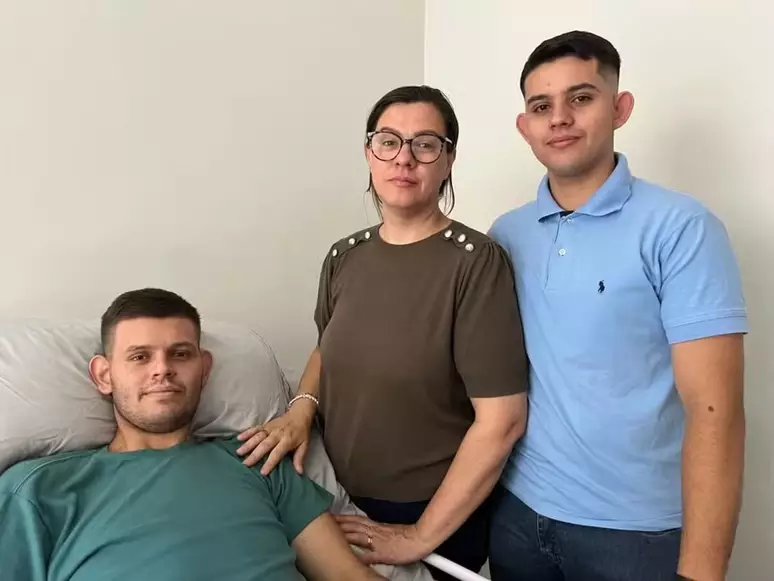 Paciente tetraplégico. Mulher e jovem abraçam homem deitado em cama, expressando apoio e união no quarto.