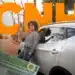 Jovem sorridente segura chave ao lado de carro branco com CNH em destaque.