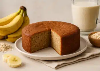 Bolo de banana caseiro com fatia cortada, bananas frescas, aveia e leite ao fundo.