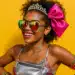 Jovem sorrindo com tiara de brilhantes, laço rosa, óculos espelhados e maquiagem com glitter.