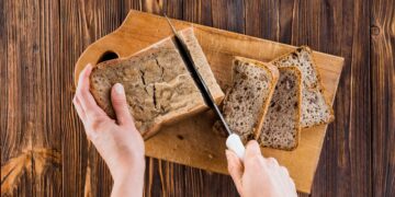 Mãos cortando pão integral em fatias sobre tábua de madeira
