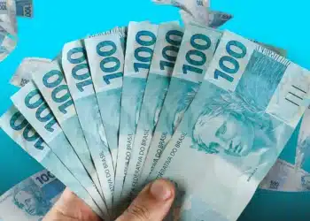 Mão exibindo leque de notas de cem reais com fundo azul vibrante
