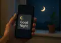 Mão segurando celular com mensagem 'Good Night' ao lado de uma janela à noite