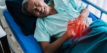 Homem idoso deitado em uma cama de hospital, expressando dor e segurando a região do abdômen com um brilho avermelhado indicando o local do desconforto.